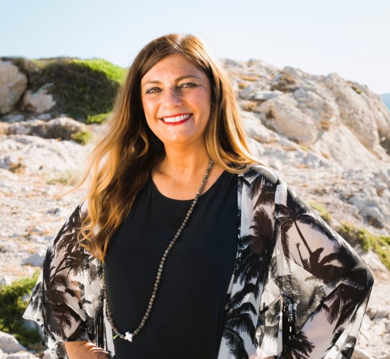 Nathalie Moraldo Femme souriante en tenue décontractée, posant sur une plage rocheuse ensoleillée.