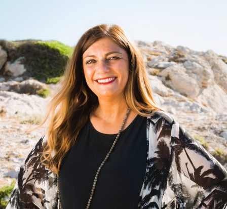 Nathalie Moraldo Sophrologue, cliente, souriante en tenue décontractée, posant sur une plage rocheuse ensoleillée.