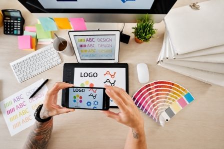 Création de marque, logo, signalétique, impression dépliant, papeterie Main d'un utilisateur prenant une photo d'un logo sur un smartphone, avec un environnement de travail.